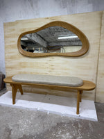 May include: A wooden bench with a light gray cushioned seat and a matching wooden mirror with an organic shape. The mirror is mounted on a light-colored wooden panel. The bench has four legs.