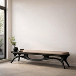 May include: A modern, dark wood bench with a beige cushion. The bench has an organic, flowing design with curved legs and a built-in side table. A small black vase with greenery and books are on the side table.