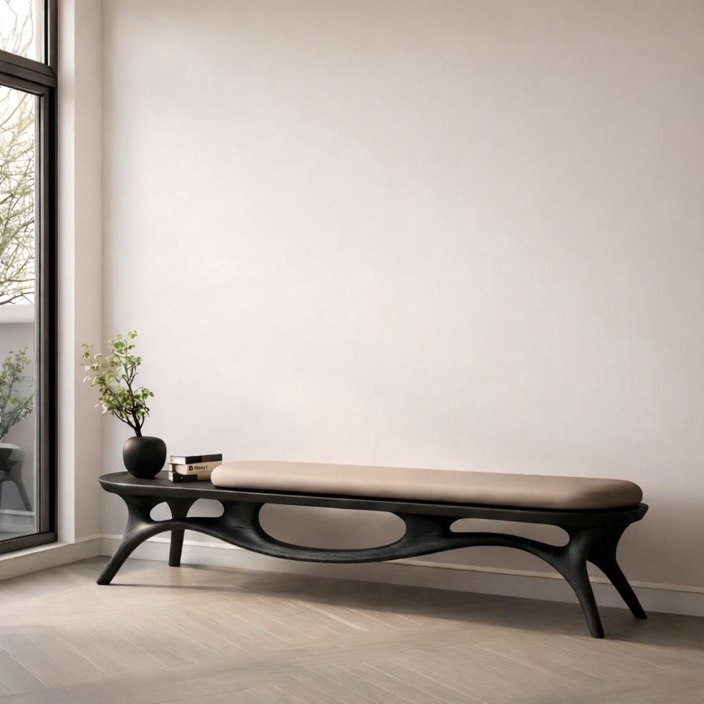 May include: A modern, dark wood bench with a beige cushion. The bench has an organic, flowing design with curved legs and a built-in side table. A small black vase with greenery and books are on the side table.