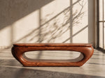 May include: A solid, wooden coffee table with a unique, curved design. The table is a warm, reddish-brown color and features an oval-shaped opening in the center. The table measures approximately 60 inches long and 20 inches wide.