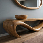 May include: A wooden console table with a unique, flowing design. The table is made of light brown wood and features a curved base. A wooden bowl sits on the tabletop. A round wooden mirror hangs on the gray wall above the table.