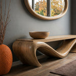 May include: A wooden console table with a unique, flowing design, featuring a large wooden bowl on top. The table has a natural wood finish and is set against a gray wall with a round mirror and a decorative vase.