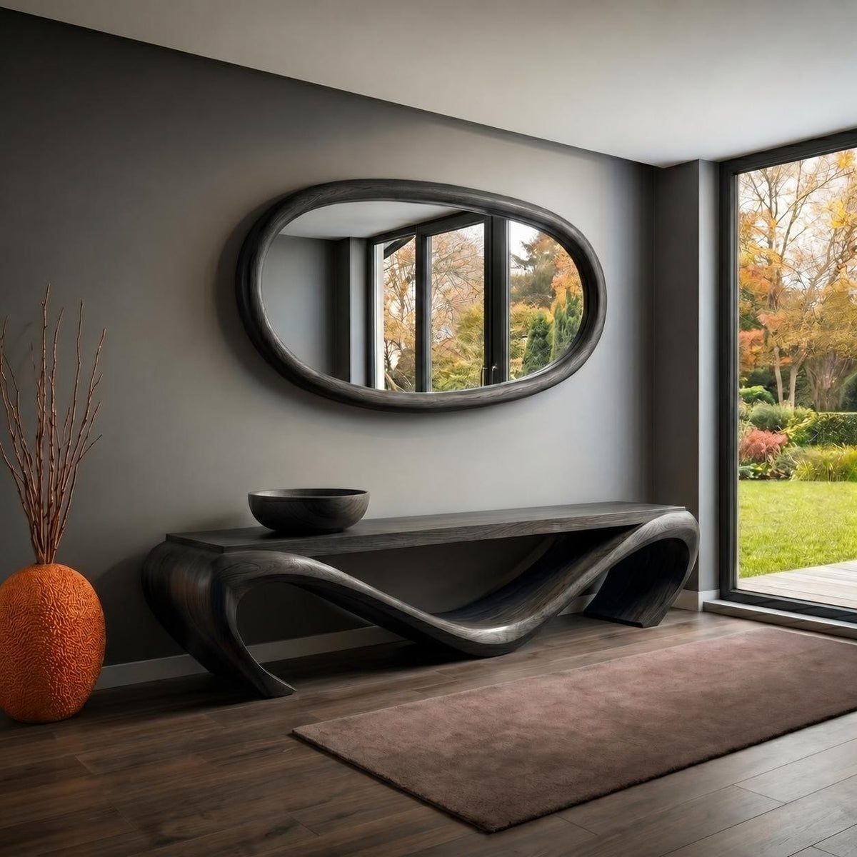 May include: A modern entryway featuring a dark wood console table with a unique, flowing design. Above the table hangs an oval mirror with a dark frame. An orange vase with dried branches and a dark bowl complete the decor. A brown rug and a large window are also visible.