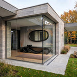 May include: Exterior view of a modern home with large glass windows. Inside, a dark console table with a unique, curved design is visible, along with a large oval mirror. The building features stone cladding and a minimalist aesthetic.