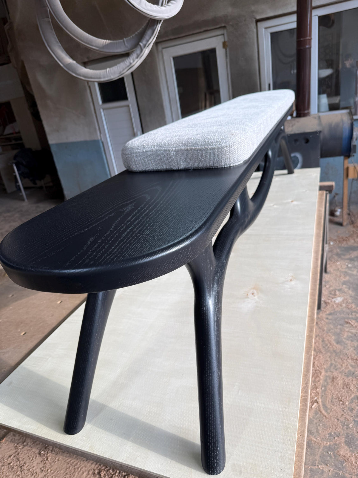 May include: A modern black wooden bench with a gray upholstered seat. The bench features a curved design with unique, branch-like legs. The wood grain is visible on the bench's surface, adding texture and visual interest to the piece.