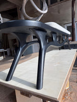 May include: A modern black wooden bench with a curved design. The bench has a light gray upholstered seat and unique, sculptural legs. The bench is placed on a light-colored wooden surface.