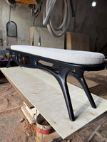 May include: A modern bench with a black wooden frame and a light gray upholstered seat. The bench has a unique, curved design with four legs. The seat appears to be approximately 60 inches long.