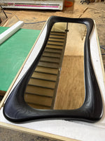 May include: A black, uniquely shaped mirror with a wooden frame. The mirror has a curved, elongated design. The reflection shows a staircase. The mirror is set in a wooden frame and rests on a green surface.