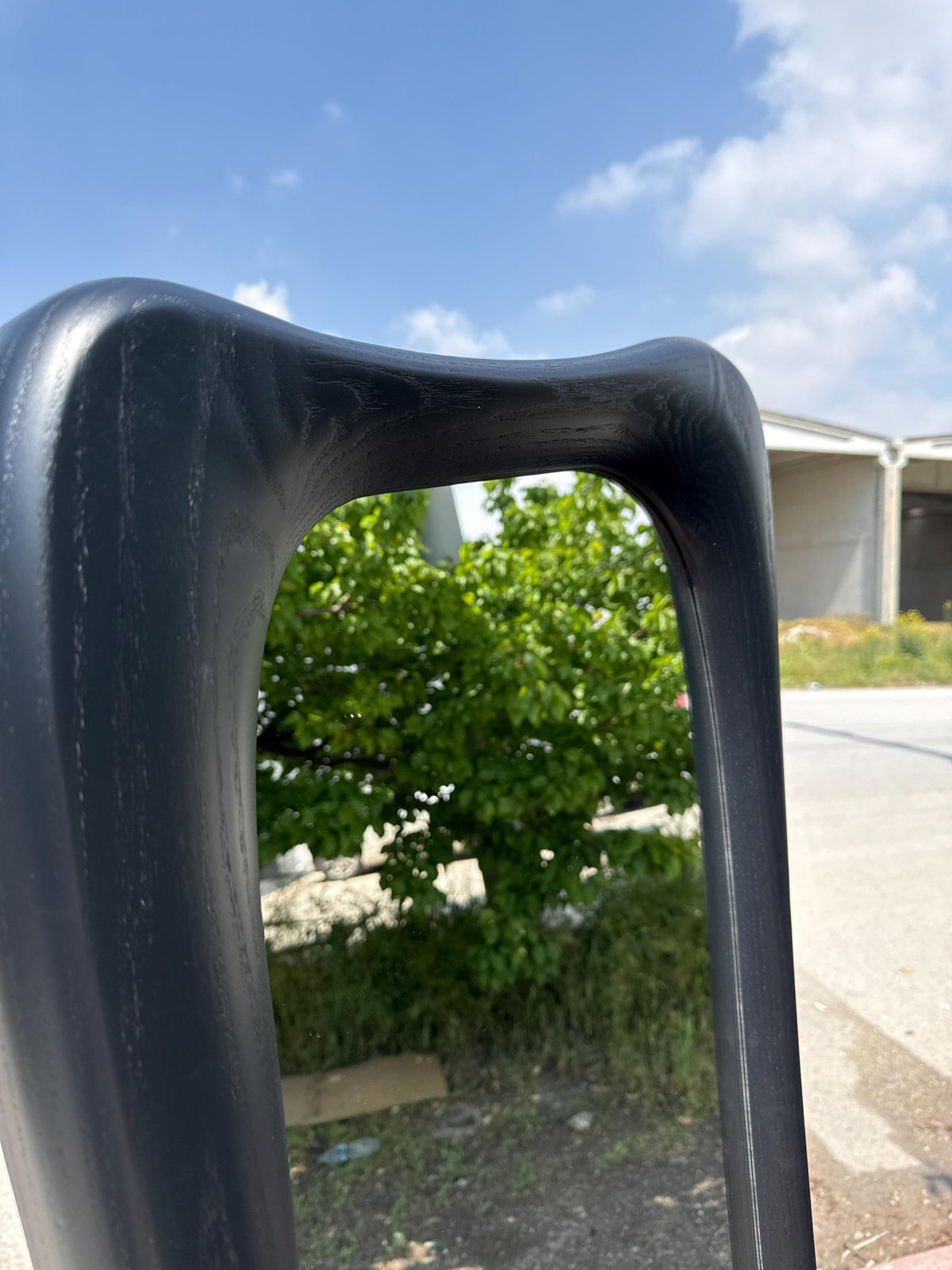 May include: A close-up of a black chair back with a curved top. The chair back is made of a dark material with a wood grain texture. The background shows green foliage and a blue sky with clouds.