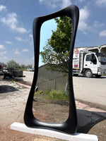 May include: A tall, black, free-standing mirror with a unique, curved frame. The mirror reflects a blue sky, trees, and a truck. The frame has a textured appearance and is supported by a white base. The mirror's shape is elongated and organic.