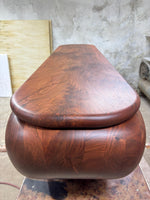 May include: A handcrafted wooden box with a rounded, oval shape and a matching lid. The box is made of dark brown wood with a visible grain pattern. The lid sits flush on top of the box.