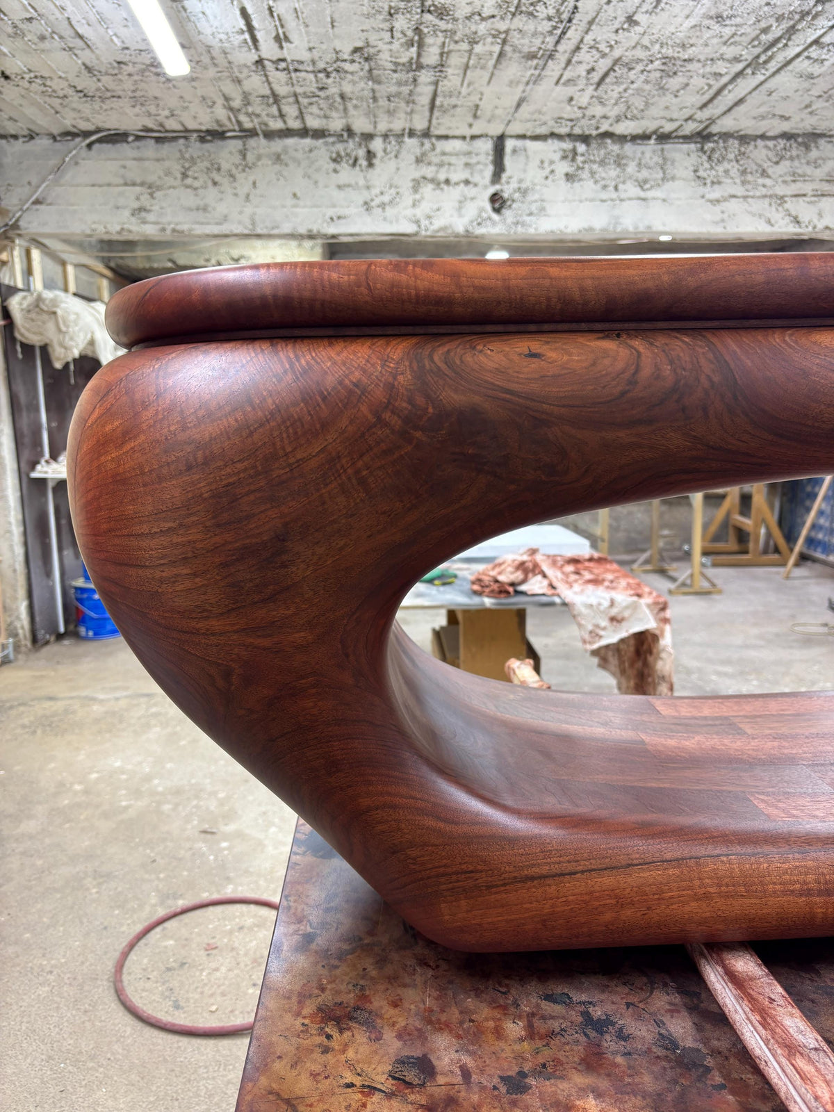 May include: A handcrafted, solid wood coffee table with a rich, reddish-brown finish. The table features a unique, curved design with a smooth, rounded top and a distinctive open space in the center. The wood grain is visible, adding to its natural aesthetic.