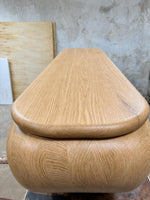 May include: A light-colored wooden storage bench with a rounded base and a flat, slightly tapered top. The wood grain is visible, and the bench appears to be newly constructed. The design is modern and minimalist.