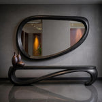 May include: A modern, dark-stained wood console table with a matching oval mirror. The table has a unique, flowing design with curved legs. Three brown vases sit on the table. The mirror reflects a room with warm lighting.