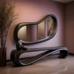 May include: A modern, dark wood console table and mirror set. The mirror has an organic, flowing shape, and the table features a curved design. Two black vases sit on the table. The room has a neutral color palette.