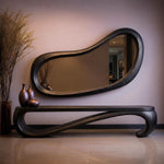 May include: A modern, dark-stained wooden console table with a matching, organically shaped mirror. The table has a curved, flowing design. Three brown vases sit on the table, and tall, dried plants are in a vase to the left.