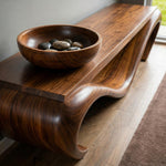 May include: A dark brown wooden console table with a curved design. A wooden bowl filled with smooth, dark stones sits on top. The table has a smooth, polished finish and a modern aesthetic.