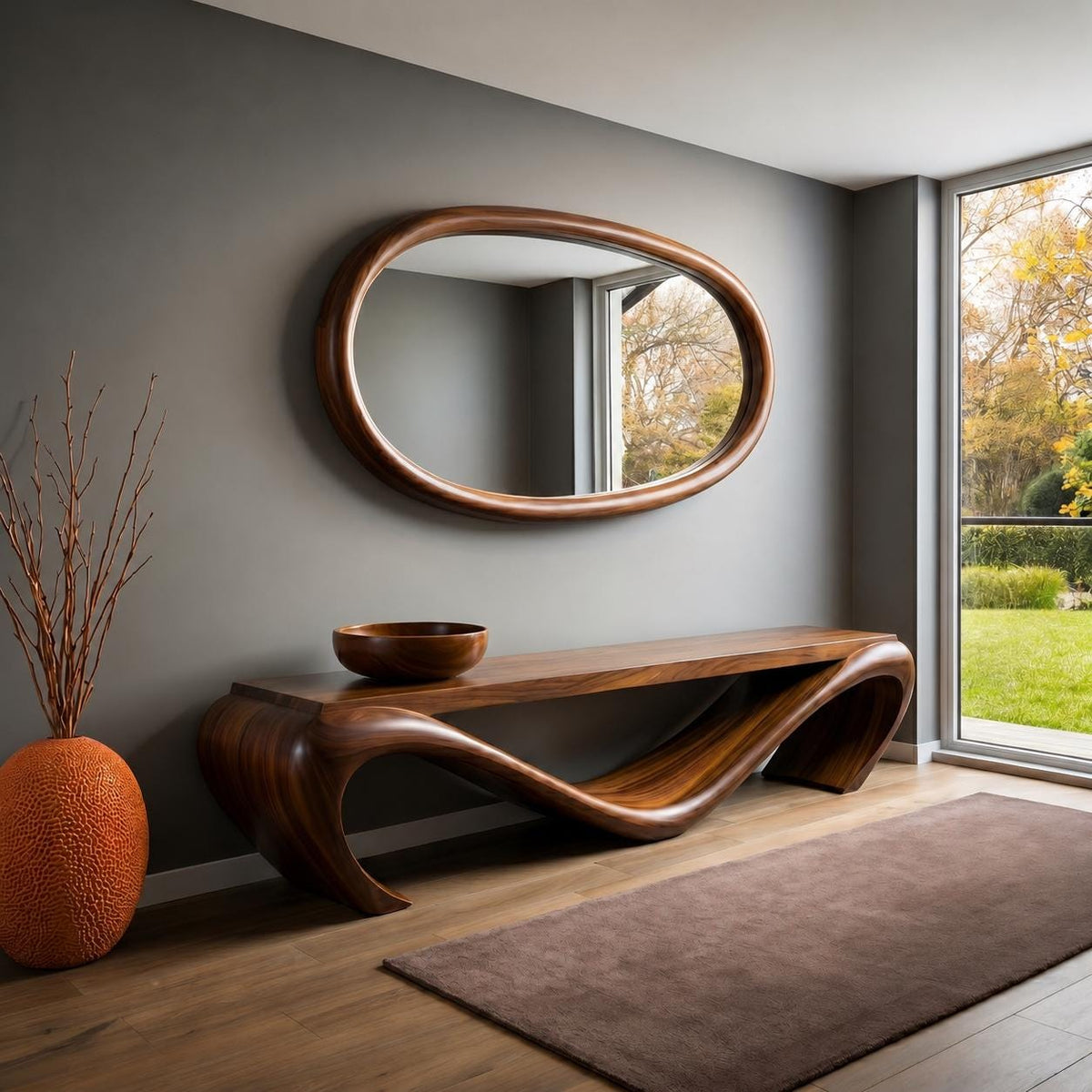 May include: A modern, wooden console table with a unique, flowing design. Above it hangs an oval-shaped mirror with a wooden frame. A wooden bowl sits on the table. An orange vase with branches and a brown rug complete the room's decor.