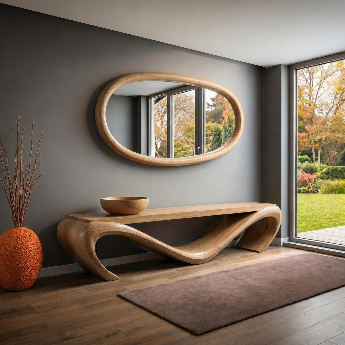 May include: A modern wooden console table with a unique, flowing design. Above it, an oval-shaped mirror with a wooden frame hangs on a gray wall. A decorative orange vase and a wooden bowl complete the decor. The room has a view of a lush garden.