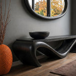 May include: A black wooden console table with a unique, flowing design. A black bowl sits on the table's surface. An orange textured vase and a round mirror are also in the image. The room has a modern, minimalist aesthetic.