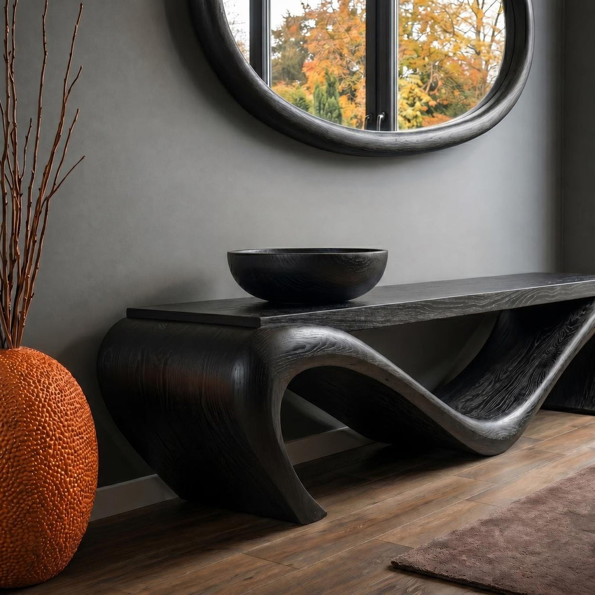 May include: A black wooden console table with a unique, flowing design. A black bowl sits on the table's surface. An orange textured vase and a round mirror are also in the image. The room has a modern, minimalist aesthetic.