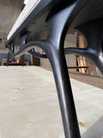 May include: Close-up of a modern bench with a black, organically shaped wooden frame. The bench has a light gray upholstered seat. The legs are curved and flared, providing a unique design element. The bench is set on a light-colored wooden surface.