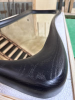 May include: Close-up of a black, curved wooden frame of a mirror. The frame has a smooth, polished finish with visible wood grain. The mirror reflects a staircase and a light-colored wall.