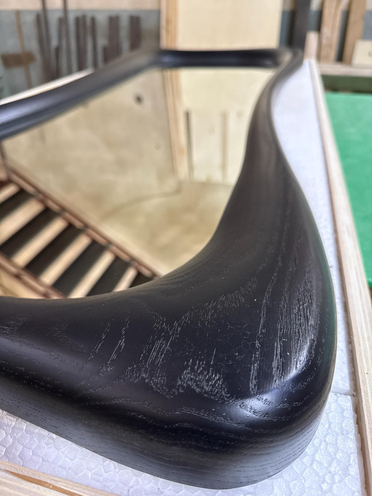 May include: Close-up of a black, curved wooden frame of a mirror. The frame has a smooth, polished finish with visible wood grain. The mirror reflects a staircase and a light-colored wall.