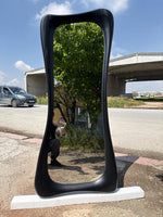 May include: A tall, black, hourglass-shaped mirror stands outdoors on a white base. The mirror's frame has a textured wood grain appearance. The reflection shows a tree and a person holding a phone. The background includes a road, a car, and a bridge.