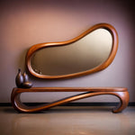 May include: A modern wooden console table with a matching, organically shaped mirror. The mirror and table are made of a warm-toned wood. Three dark brown decorative vases sit on the table.