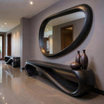 May include: A modern entryway featuring a large, oval-shaped mirror with a dark wood frame. Below the mirror is a matching dark wood console table with a curved design. Three brown vases sit on the table. A matching bench is in the background.