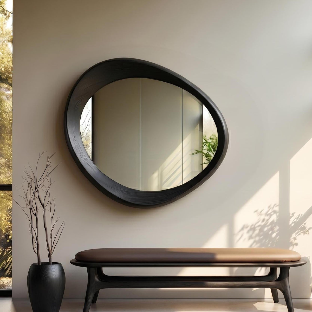 May include: A wall-mounted mirror with an asymmetrical, organic, pebble-shaped black frame. The mirror reflects a room interior. Below it sits a long, low bench with a dark brown upholstered seat and a black wooden base, plus a black vase.