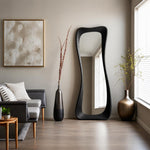May include: A tall, floor-standing mirror with a thick, black, wavy, organic-shaped frame. The mirror is positioned upright against a neutral-colored wall, reflecting the interior space, including a window with white blinds and light-colored walls.