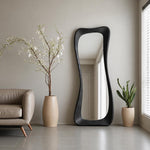 May include: A tall, floor-standing mirror with a thick, black, irregular, wavy wooden frame. The mirror has a smooth, reflective surface and is positioned upright against a plain wall, reflecting the interior space and light from a nearby window.