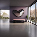 May include: A heart-shaped wall mirror with a thick black frame mounted on a dark purple wall above a low black bench featuring a soft pink cushion. The bench has a hollow center and sits on a smooth gray floor in a room with large glass windows.