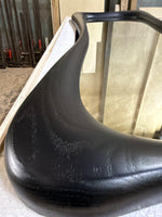 May include: A large, black-stained wooden mirror frame with a smooth, organic, and curved silhouette. The wood grain is visible through the dark finish, highlighting the natural texture of the material along the rounded, thick edges of the frame.