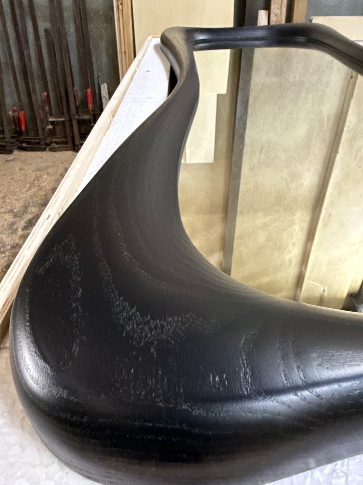 May include: A large, black-stained wooden mirror frame with a smooth, organic, and curved silhouette. The wood grain is visible through the dark finish, highlighting the natural texture of the material along the rounded, thick edges of the frame.