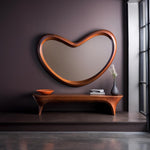 May include: A large, kidney-shaped wall mirror with a smooth, polished wooden frame. Below the mirror sits a long, low wooden bench with curved legs, holding an orange bowl, a stack of books, and a dark gray vase with white flowers.