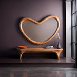 May include: A large, heart-shaped wall mirror with a thick, light-toned wooden frame. Below the mirror sits a long, curved wooden bench with four legs. On the bench are an orange bowl, a small stack of books, and a dark gray vase with dried flowers.