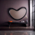 May include: A large, black, kidney-shaped wall mirror with a thick wooden frame. Below the mirror sits a long, black, curved wooden bench. On the bench are a bright orange bowl, a stack of books, and a dark gray vase with dried white flowers.
