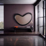 May include: A large, black, bean-shaped wall mirror hangs above a long, black, curved-leg bench. On the bench sits a bright orange bowl, a small stack of books, and a dark gray vase containing a dried floral stem against a dark purple wall.