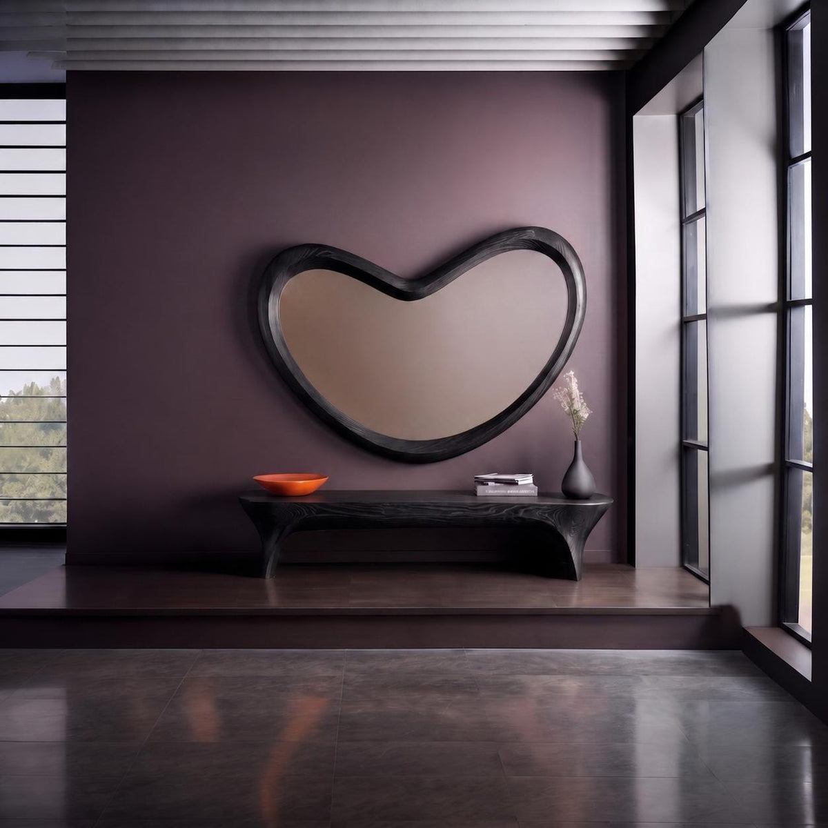 May include: A large, black, bean-shaped wall mirror hangs above a long, black, curved-leg bench. On the bench sits a bright orange bowl, a small stack of books, and a dark gray vase containing a dried floral stem against a dark purple wall.